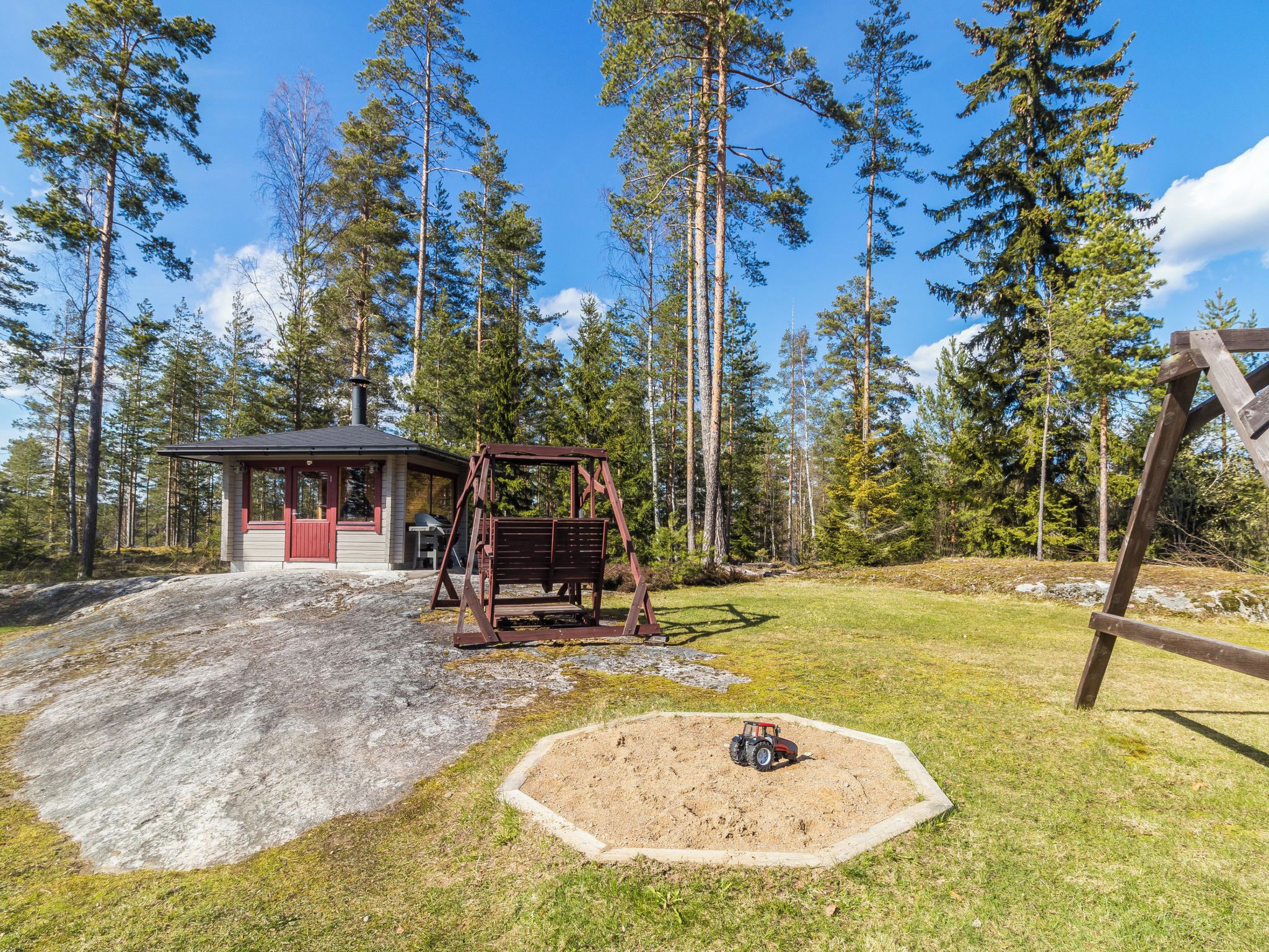 Eine Holzhütte mit Schaukel und Kiesfläche, umgeben von hohen Bäumen.