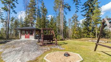 Eine Holzhütte mit Schaukel und Kiesfläche, umgeben von hohen Bäumen.
