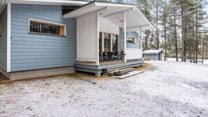 Eine moderne Holzhütte mit einer überdachten Veranda, einem Essbereich und einem kleinen Geräteschuppen in bewaldeter Landschaft.