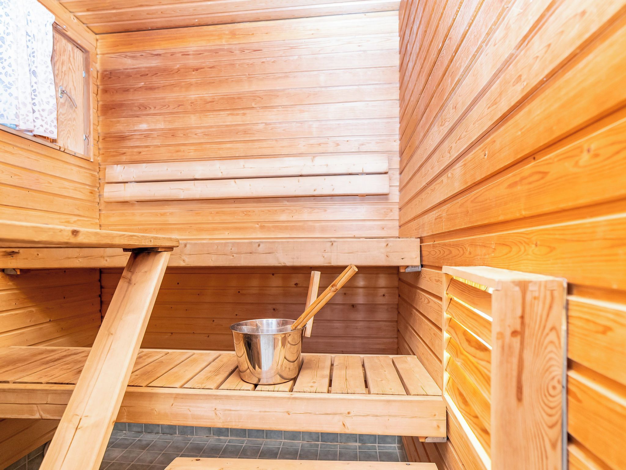 A sauna with wooden walls and benches, a metal bucket with sticks, and a tiled floor.