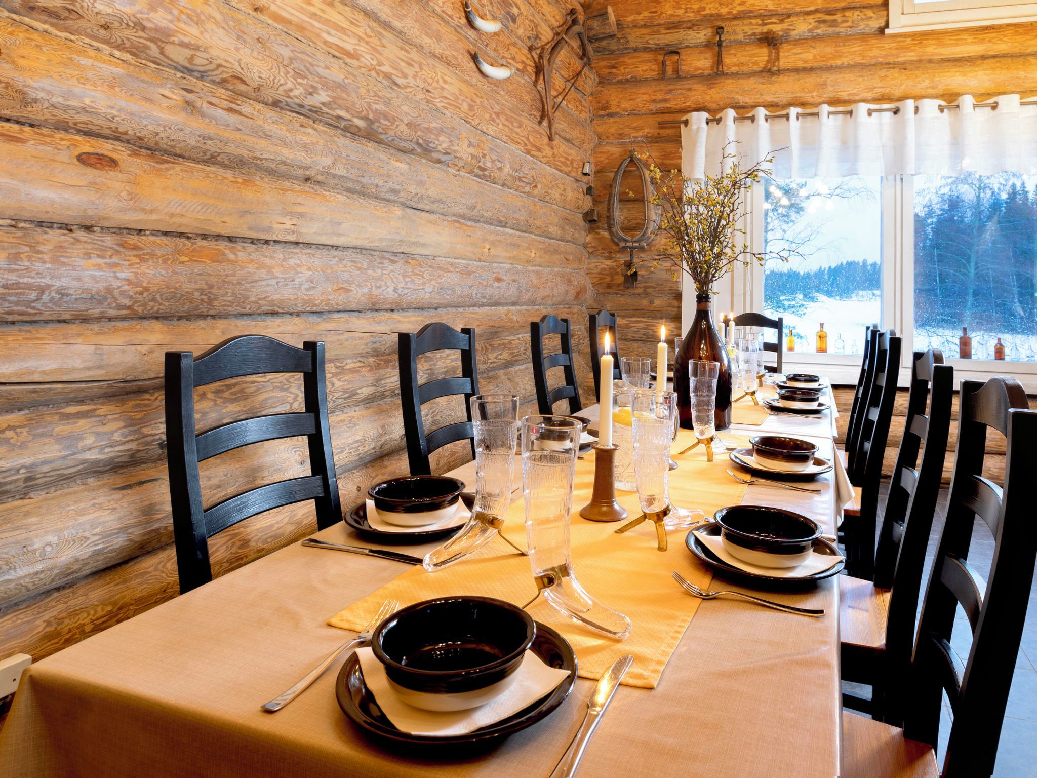 Un intérieur en bois avec une table à manger dressée pour quatre personnes, agrémentée de chaises noires, de verres transparents et d’un vase avec des branches.