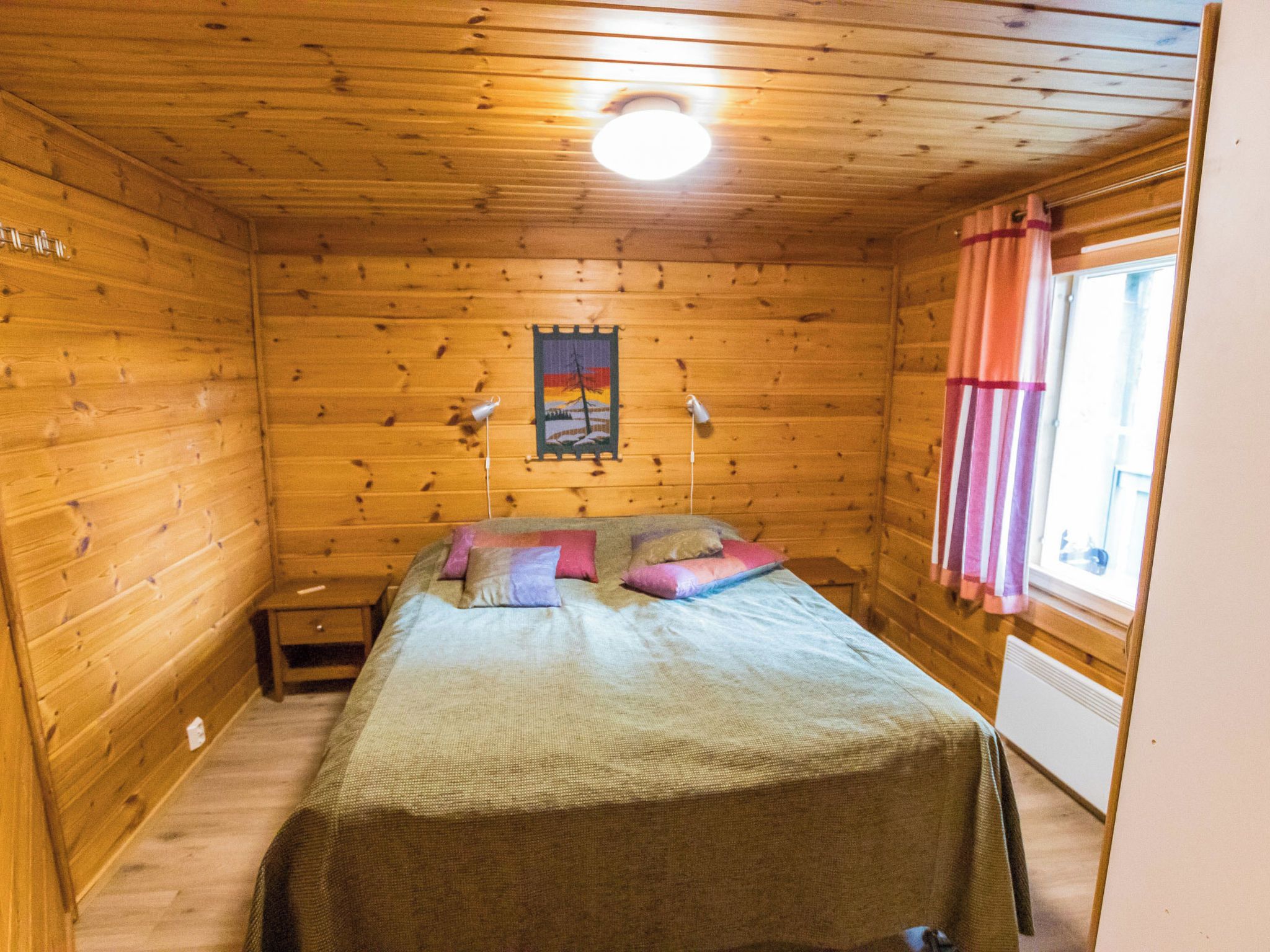A wooden cabin room with a bed, a nightstand, a wall-mounted lamp, and a window with curtains.