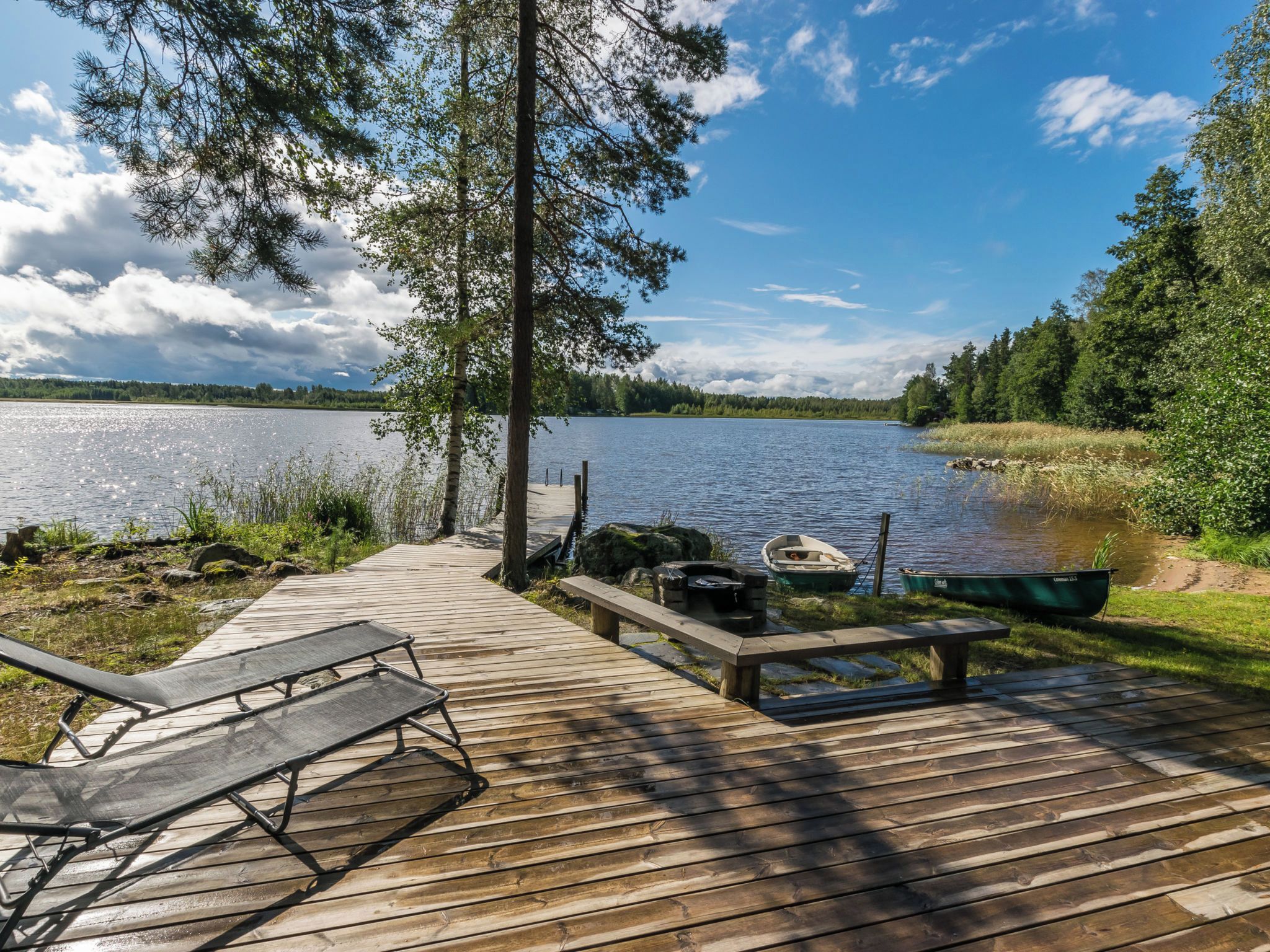 A wooden dock leading to a calm lake with boats, surrounded by trees and greenery.