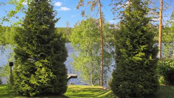 A lakeside view with two prominent evergreen trees, a grassy area, and a dock extending into the water.