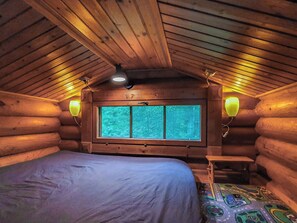 A log cabin bedroom with a bed, a window with a view, and two wall-mounted lamps.