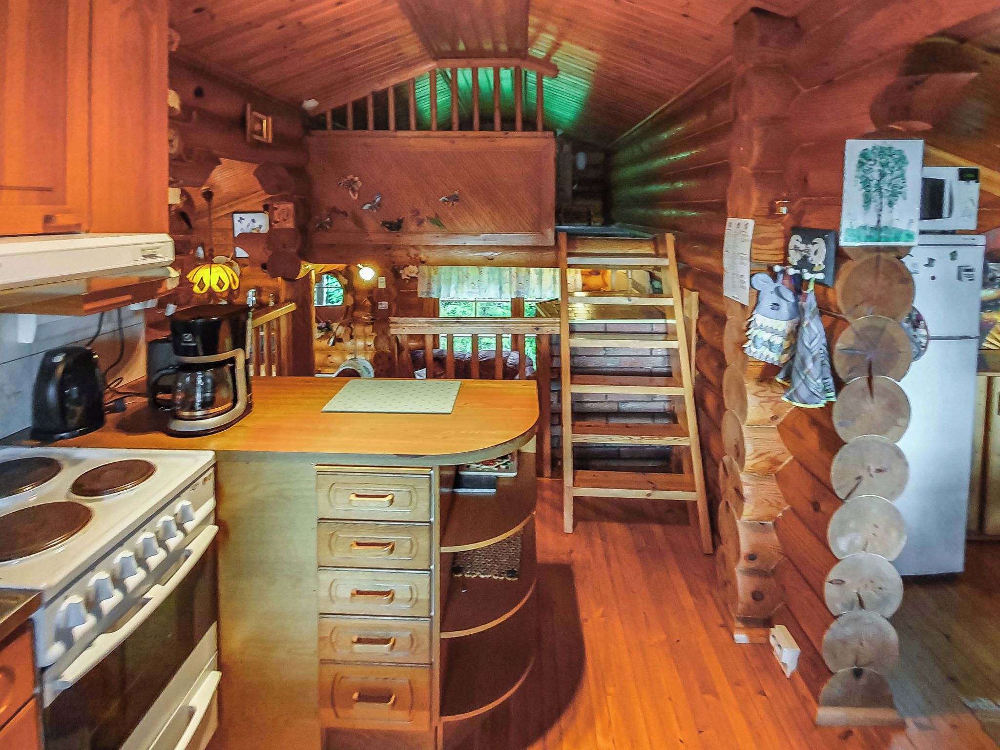 Interior rústico de una cabaña con una mesa de comedor de madera, una cocina con estufa y armarios, una escalera y un refrigerador.