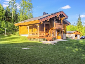 A wooden cabin with a porch and a chimney, surrounded by a grassy yard and trees.