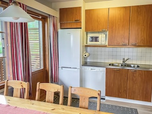 Cozinha compacta com armários de madeira, geladeira branca, micro-ondas e lava-louças. Há duas cadeiras de madeira em uma mesa.