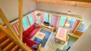 Amplia sala de estar con un sofá rojo, mesas de madera y un televisor. Grandes ventanas con cortinas ofrecen luz natural y vistas a la vegetación exterior.