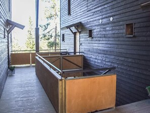 Un espacio exterior moderno con un banco de madera y una mesa con cubierta de vidrio, junto a un edificio con fachada de madera oscura y barandilla de metal.
