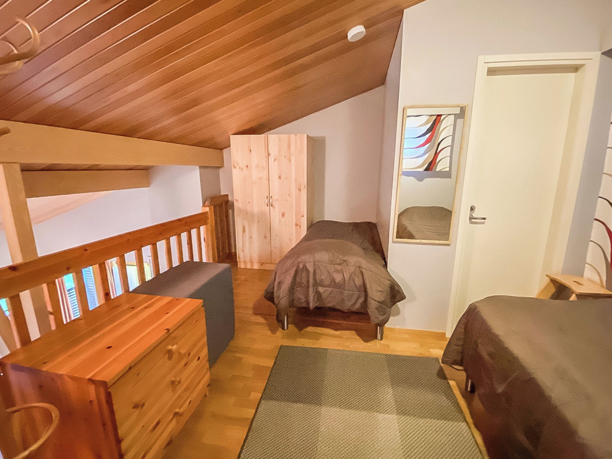 A bedroom with a wooden ceiling, a bunk bed, a single bed, a wooden bench, and a door with a window.
