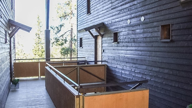 Un área de estar exterior moderna con un banco de madera y una mesa de metal, rodeada por una terraza de madera y un edificio con ventanas amplias.