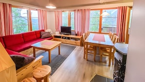 Una habitación acogedora con un sofá rojo, una mesa de comedor de madera y un televisor. Amplias ventanas con cortinas dejan entrar luz natural y ofrecen vistas al verdor exterior.