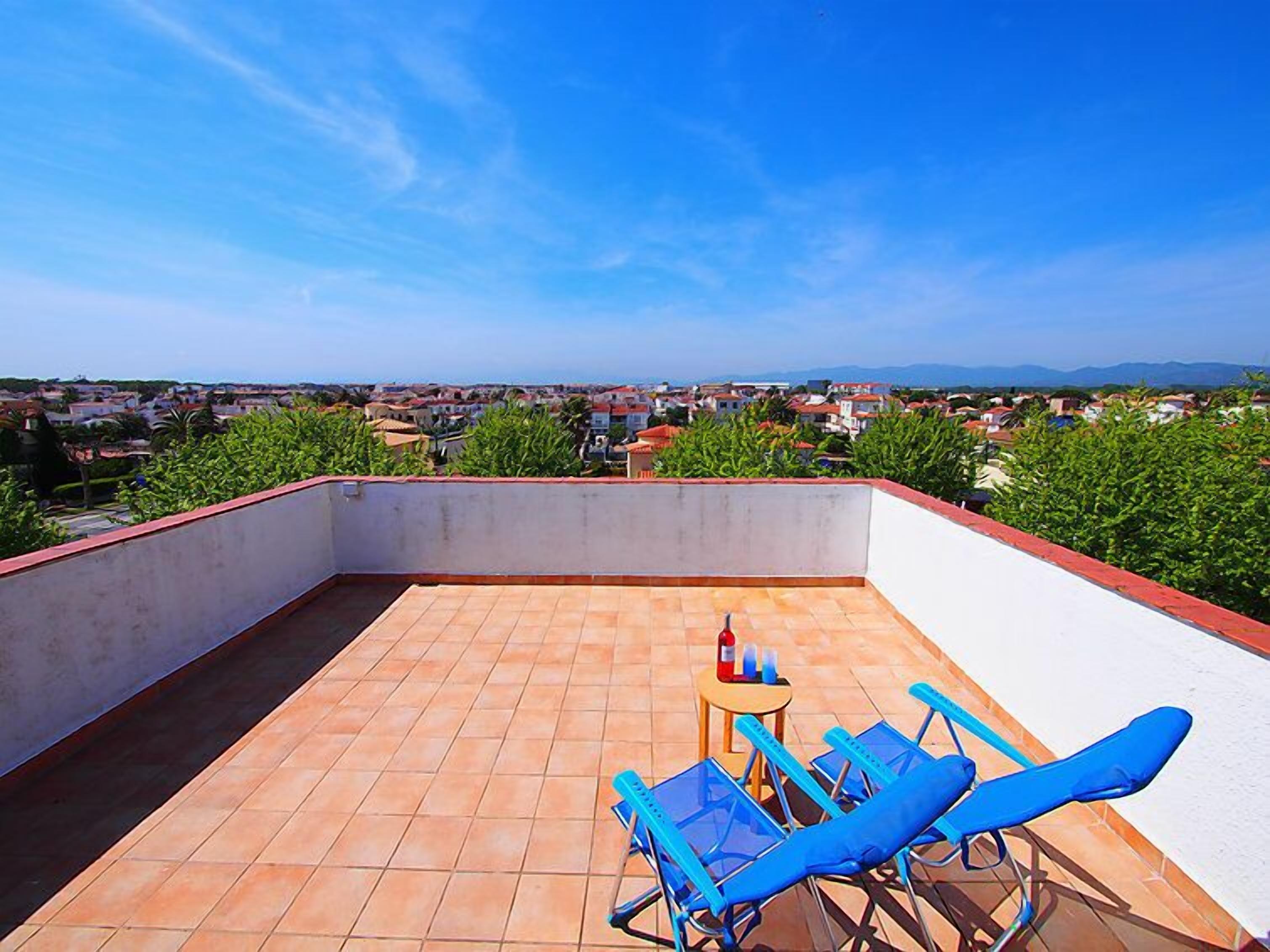 A rooftop terrace with a clear view of the city, a small table with a bottle, and blue lounge chairs.