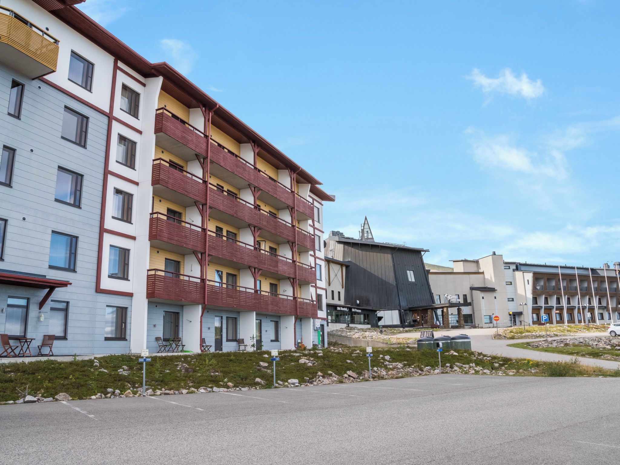 A multi-story residential building with balconies, a parking area, and a ski resort in the background.