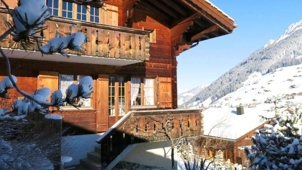 Un chalet en bois doté d’un balcon, entouré d’arbres et de montagnes couverts de neige.