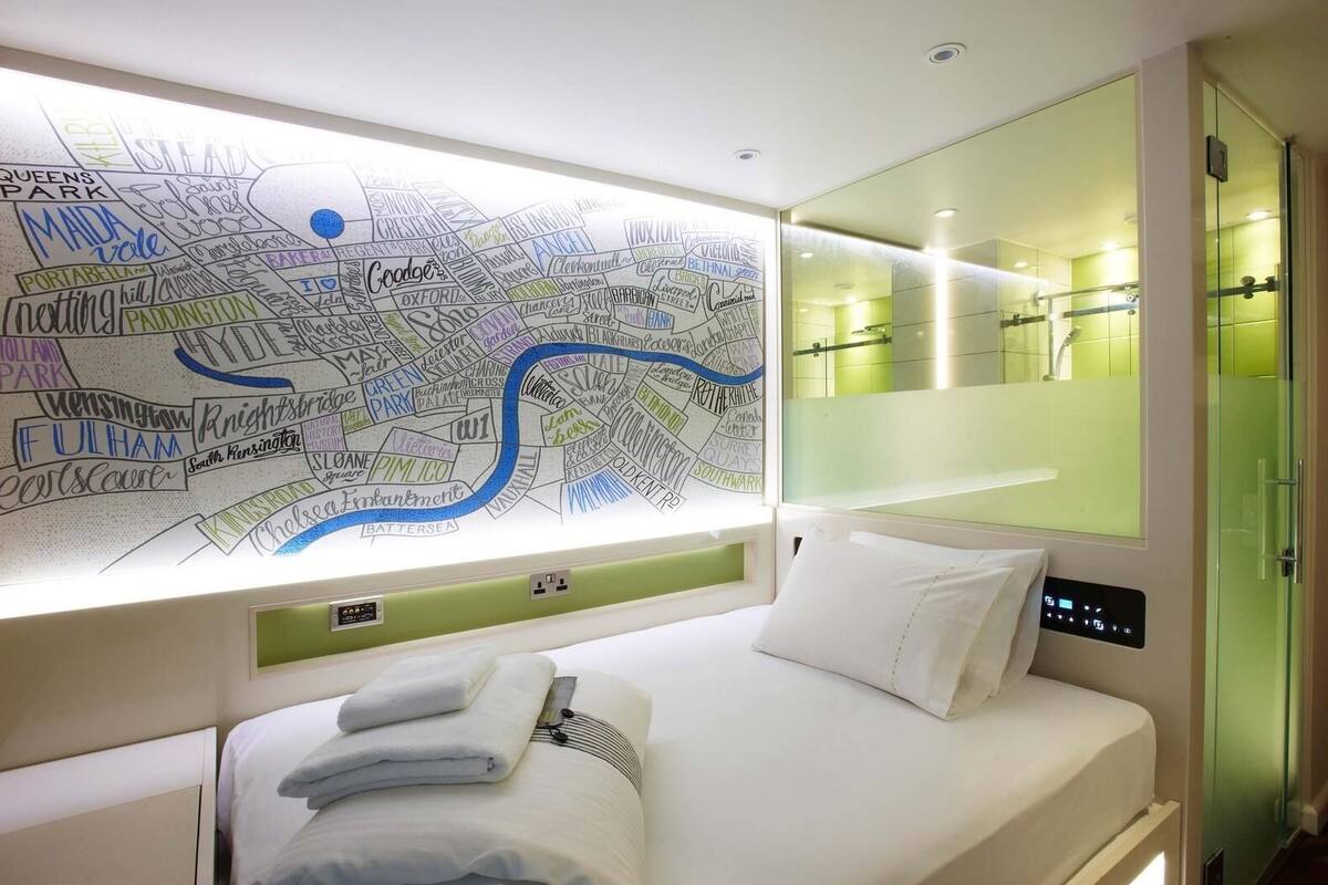 A hotel room with a large wall map, a bed with white linens, a control panel, and a glass shower enclosure.
