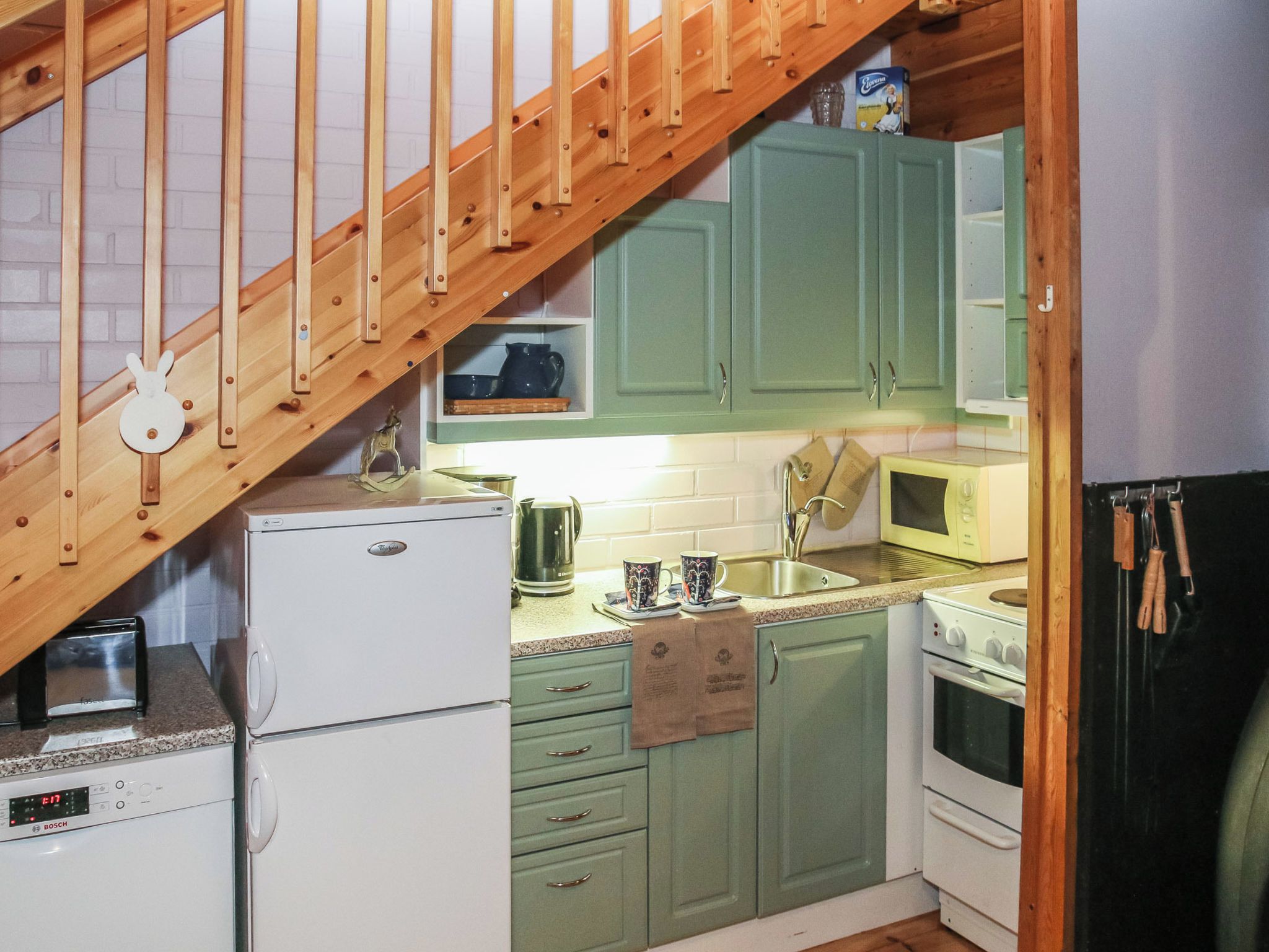 Une cuisine compacte avec un escalier en bois, un réfrigérateur Bosch, un four à micro-ondes et des armoires vertes.