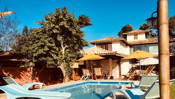 Zona de piscina con sillones de descanso y sombrillas, rodeada por un edificio con tejado de baldosas y un árbol grande.
