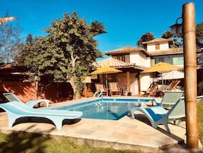 A pool area with lounge chairs and umbrellas, surrounded by a building with a tiled roof and a large tree. - Pousada L'Grace Ferradura (Búzios)