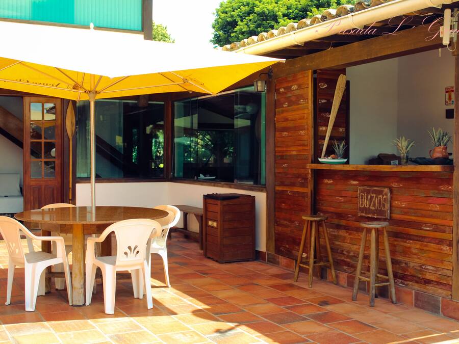An outdoor patio with a wooden table and white plastic chairs, a yellow umbrella, and a wooden bar counter with stools.