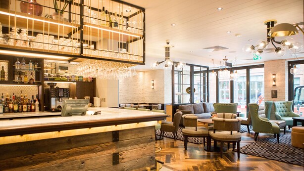 Zona de bar con mostrador de madera, asientos con sillas acolchadas y techo alto con luces colgantes.