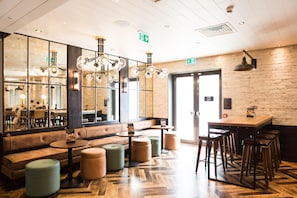 A modern interior with a wooden floor, a seating area with a bench and stools, a bar counter with stools, and pendant lighting. - Hub By Premier Inn London King's Cross (London)