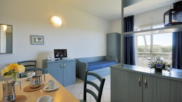 A room with a dining table, chairs, a small kitchenette, a bed, and a window with curtains. - Villaggio Turistico Residence La Cecinella (Cecina)
