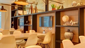A modern conference room with a large table, white chairs, a flat-screen TV, and decorative items on shelves. - hub by Premier Inn London Tottenham Court Road (Goodge Street) (London)