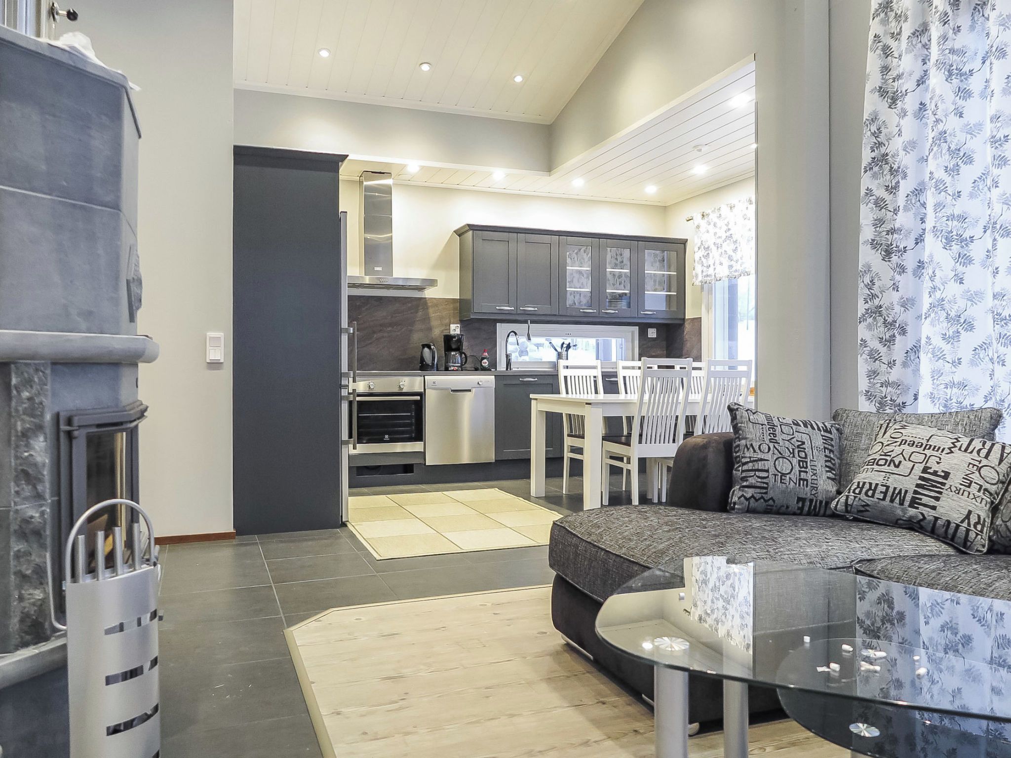 A modern living room with a glass coffee table, a sofa with patterned cushions, and a view into the kitchen area.