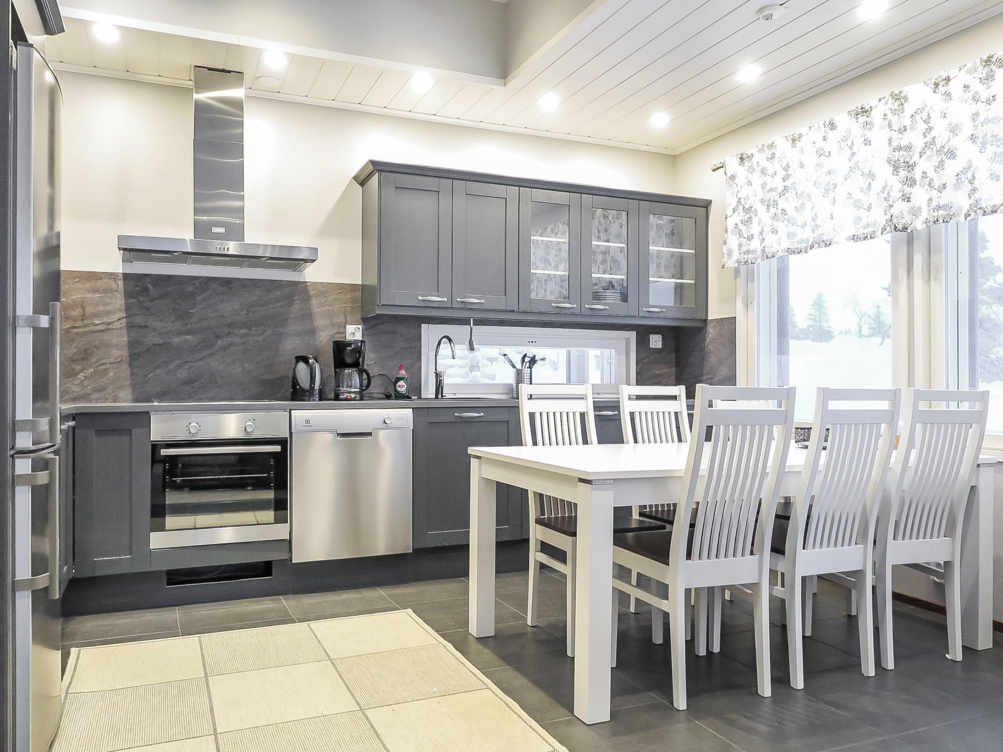 Een moderne keuken met grijze kastjes, een ingebouwde oven en een eethoek met witte stoelen en een tafel.