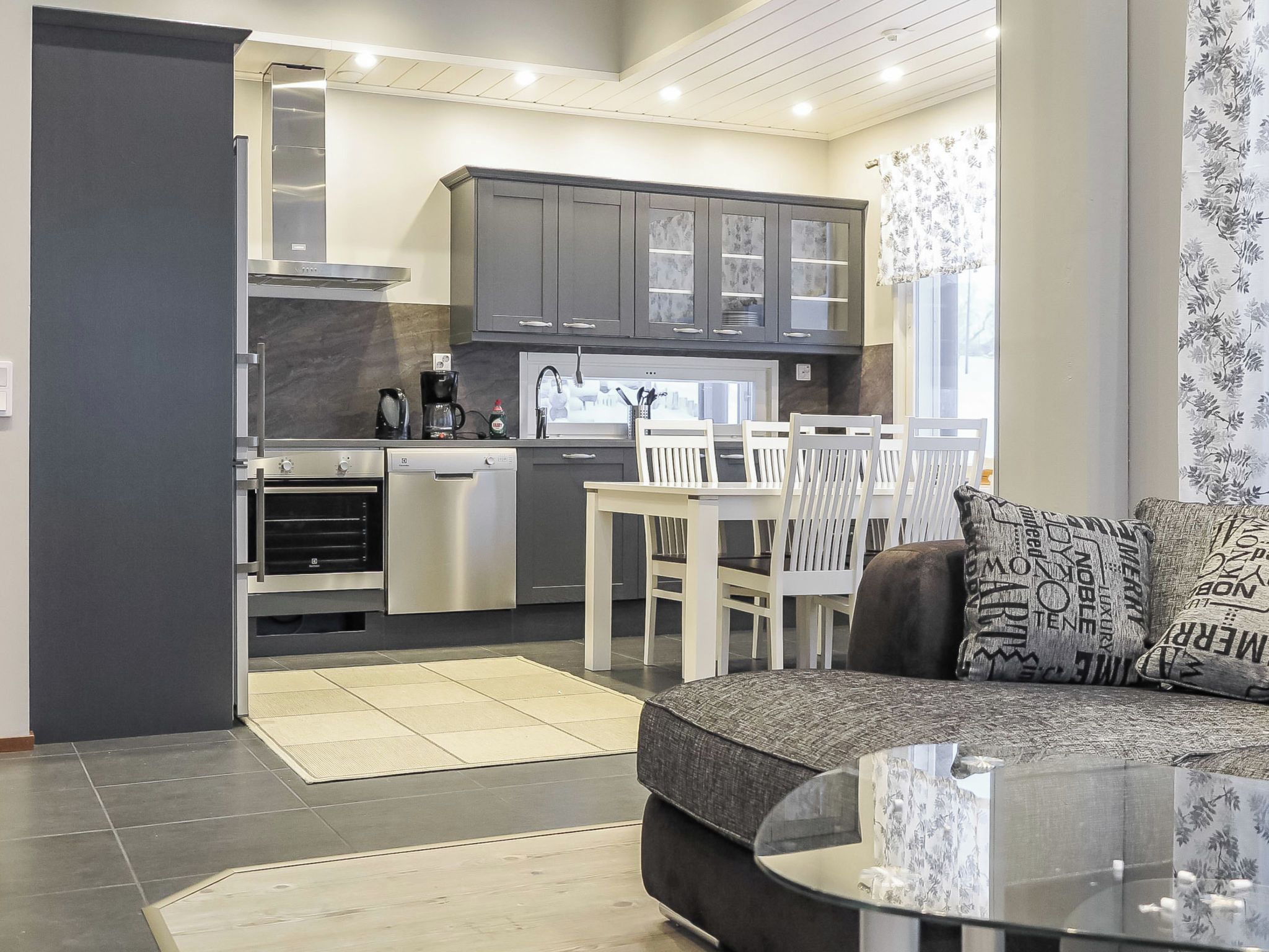 Una cocina moderna con armarios oscuros, una zona de comedor con mesa y sillas blancas, y una sala de estar con sofá oscuro y cojines estampados.