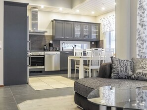 Een moderne keuken met donkere kasten, een zithoek met een witte tafel en stoelen, en een woonkamer met een donkere bank en gekleurde kussens.