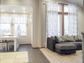 Een woonkamer met een hoekbank, een zithoek met een tafel en stoelen, en een groot raam met bloemen gordijnen.