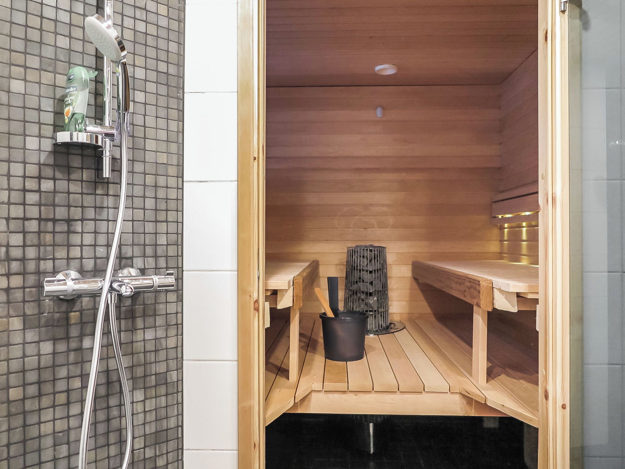 Een sauna met houten banken, een zwarte emmer en een doucheruimte met een betegelde wand en een fles biologische zeep.