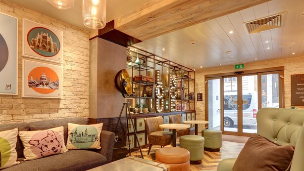 Eine Lobby mit einer Couch, Poufs, einer Bar mit Spiegel und gerahmten Bildern an der Wand.