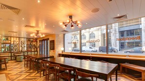 Ein Restaurantinneres mit Holztischen, Stühlen, einem Fischgrätparkett und großen Fenstern mit Blick auf die Stadt.