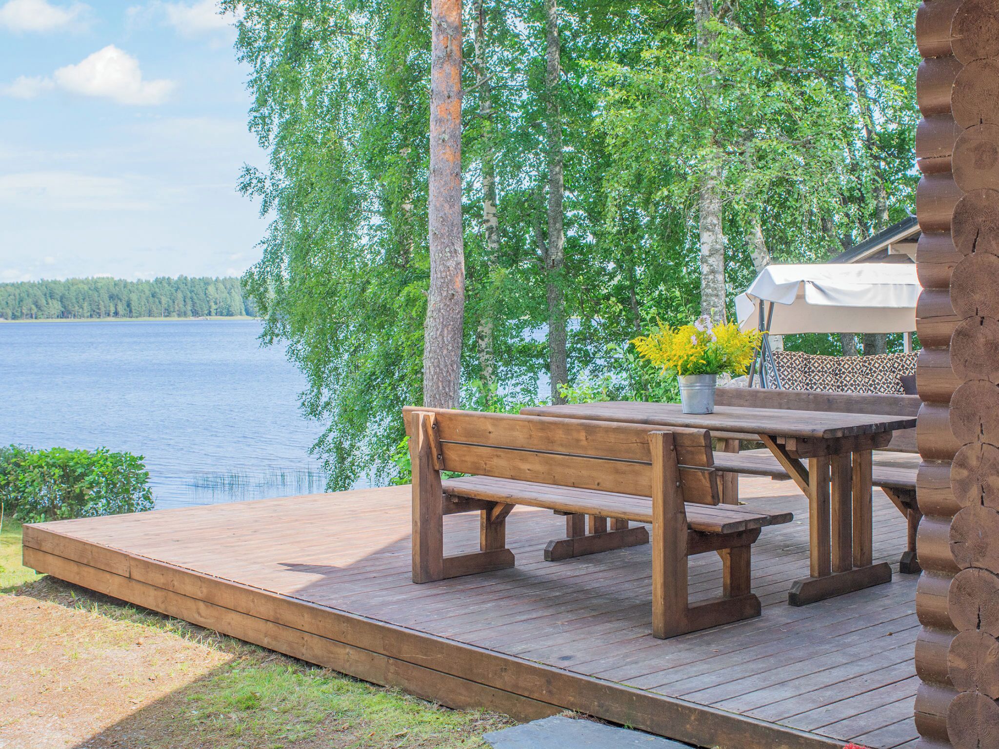 A wooden deck with a bench and picnic table, overlooking a lake with trees in the background.