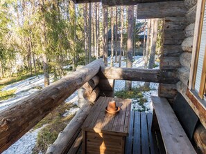 Une terrasse en bois avec une structure de cabane rustique en rondins, un chapeau posé sur une table et un paysage de forêt enneigée en arrière-plan.