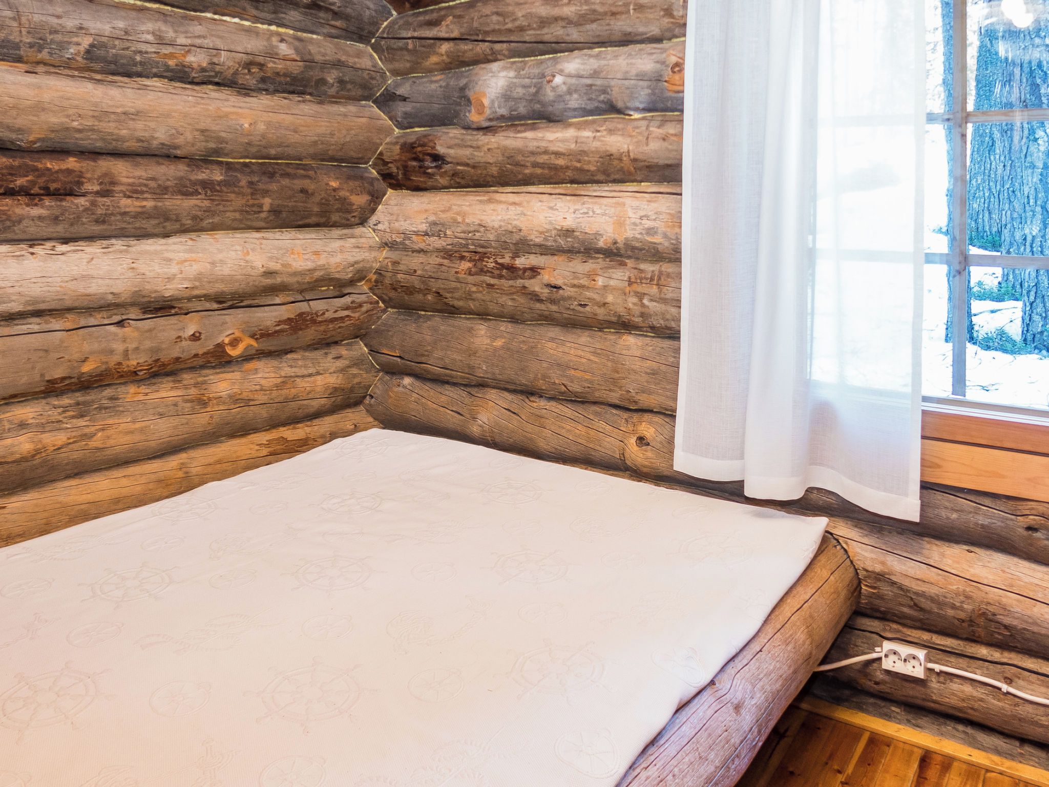 Una habitación tipo cabaña de madera con una cama, una ventana que da a los árboles y un enchufe en la pared.