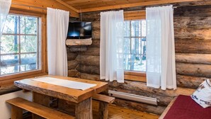 A rustic cabin interior with a wooden table and benches, a flat-screen TV mounted on a stone wall, and large windows with white curtains. - Retkietappi honkapirtti by Interhome (Kuusamo)
