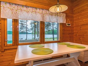 A wooden cabin with a table and four green placemats, a hanging light fixture, and a view of a lake through large windows.