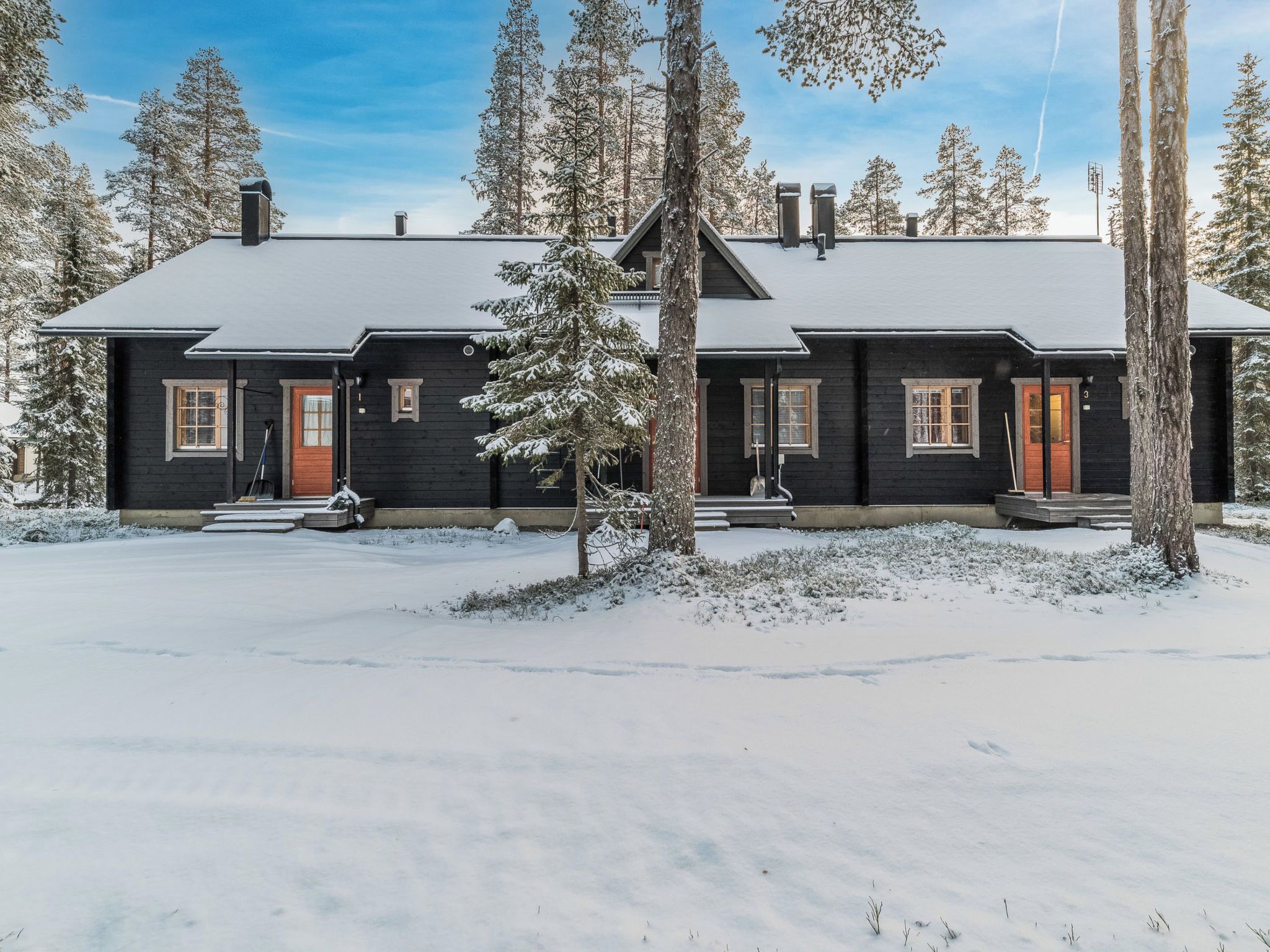 Kaksikerroksinen hirsihökkelö, jonka ulkoseinät ovat tummat, etuovi punainen ja maisema lumen peitossa.