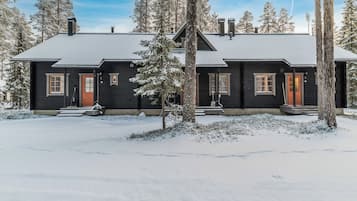 Kaksikerroksinen hirsihökkelö, jonka ulkoseinät ovat tummat, etuovi punainen ja maisema lumen peitossa.