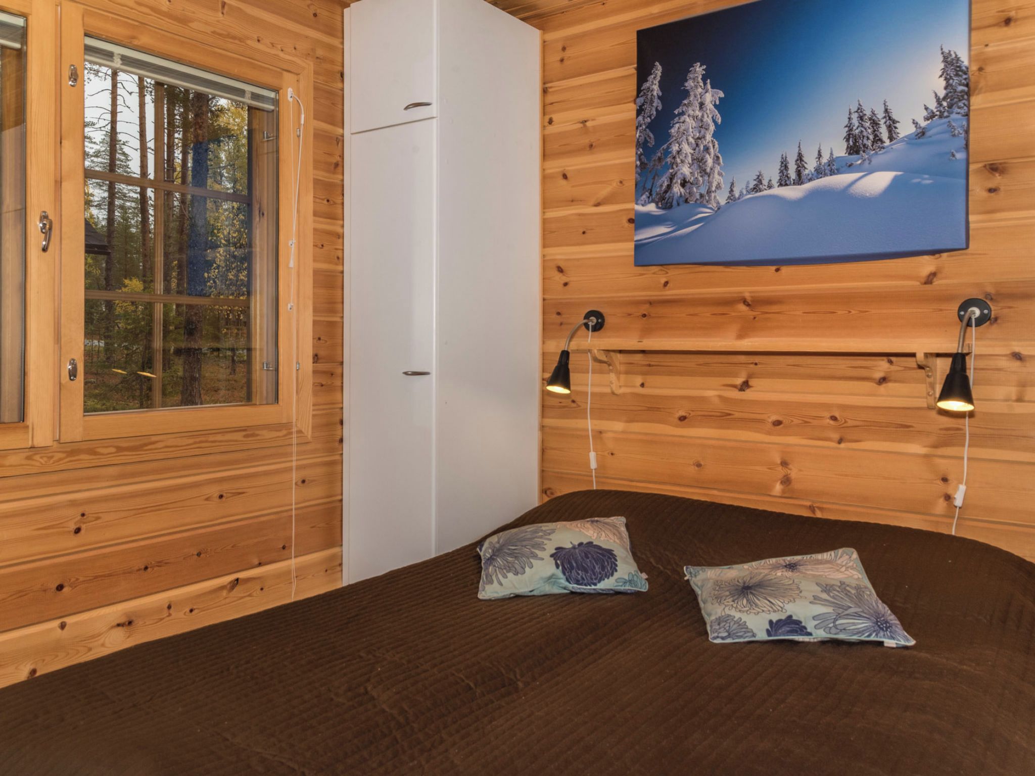 Una habitación tipo cabaña de madera con una cama, un ventanal, un armario blanco y un cuadro de un paisaje nevado.