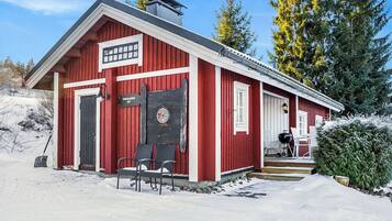 Punainen puinen rakennus, jossa on savupiippu, ovi ja kaksi tuolia sen ulkopuolella.