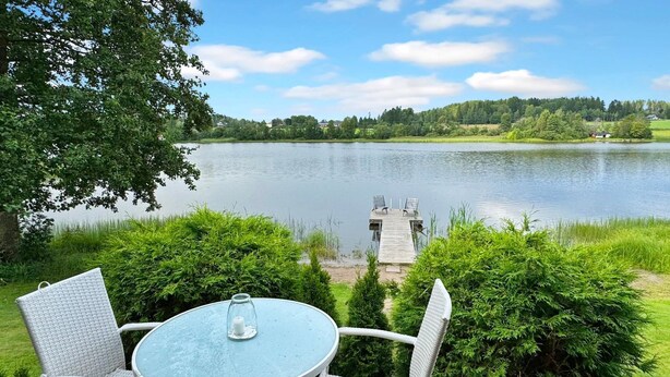 Eine Terrasse mit Korbstühlen und einem Tisch mit Blick auf einen See, in der Ferne ein Steg.