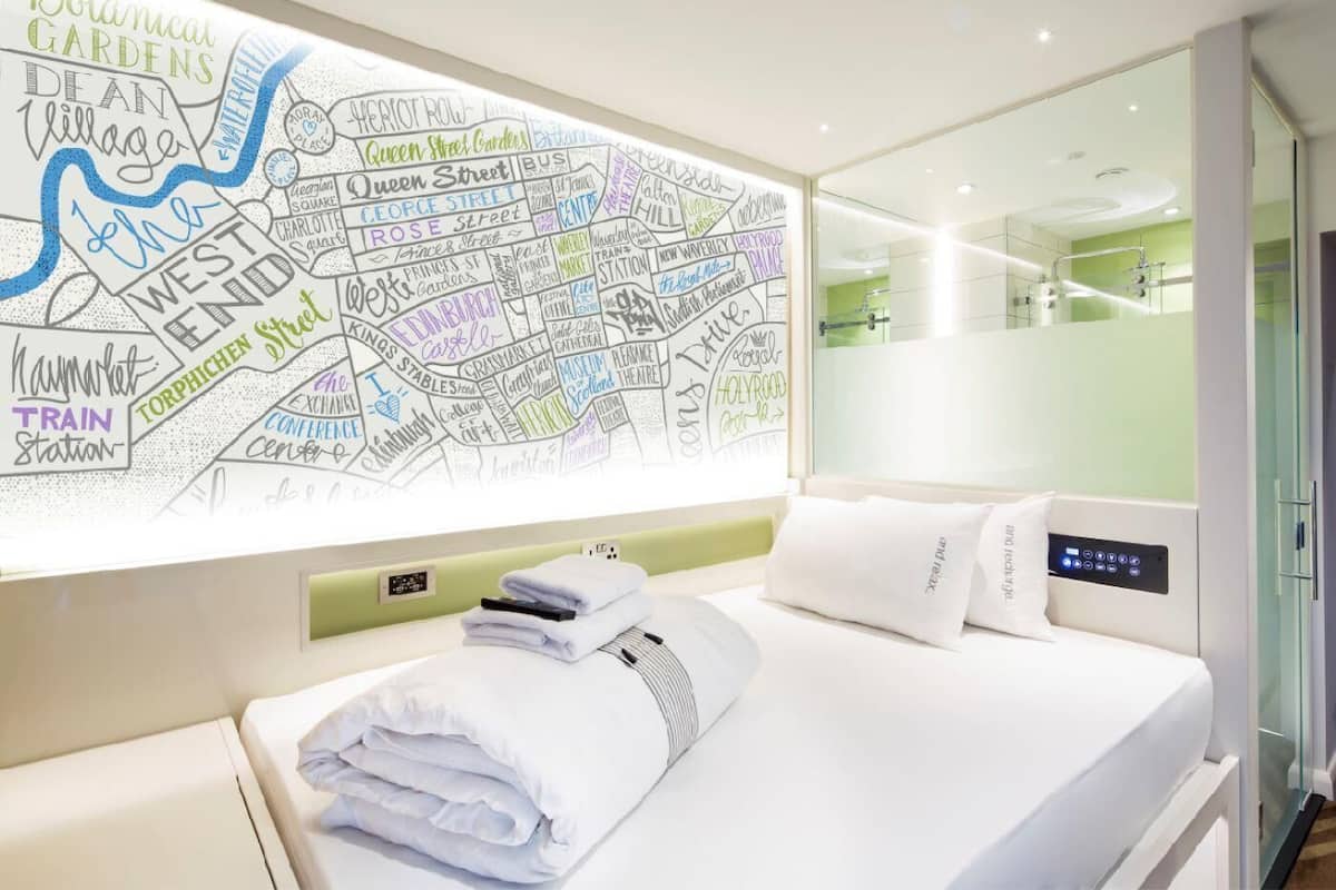 Quarto de hotel com um grande mapa na parede, cama bem arrumada, mesa de cabeceira com telefone e um livro, e um banheiro moderno visível através de um painel de vidro.