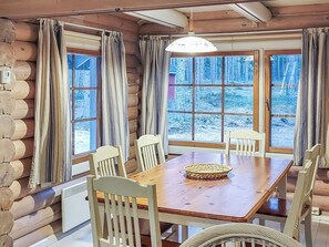 A log cabin interior with a wooden dining table, chairs, and a large window with curtains.