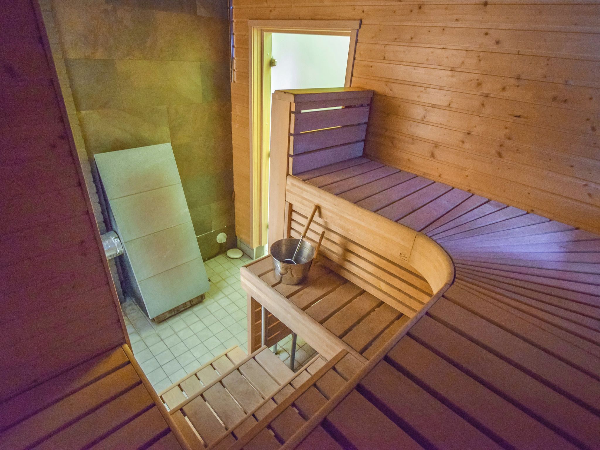 A small, compact room with a wooden bench, a metal bucket, and a window.